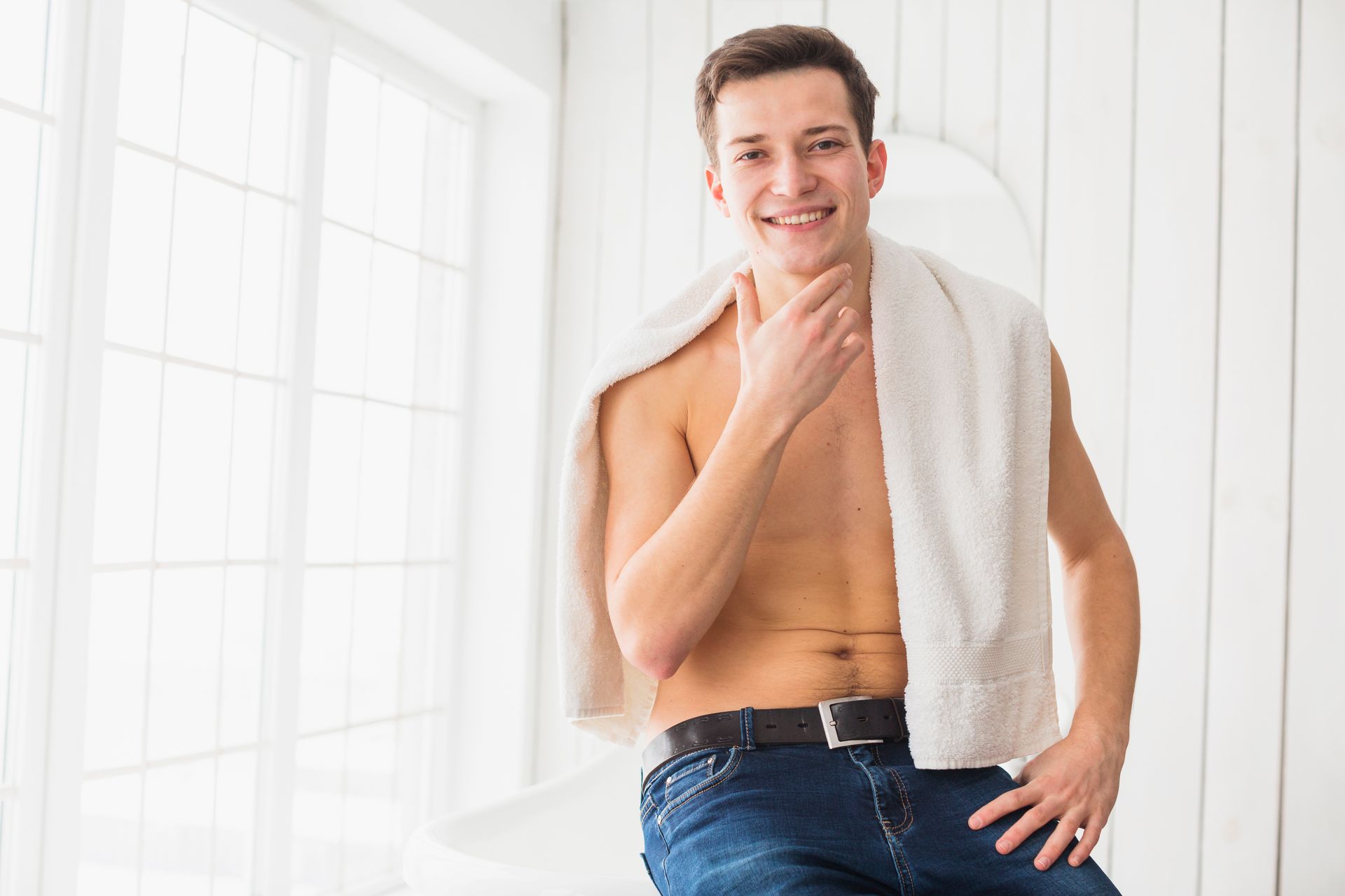 A shirtless man is sitting on a bed with a towel around his neck.