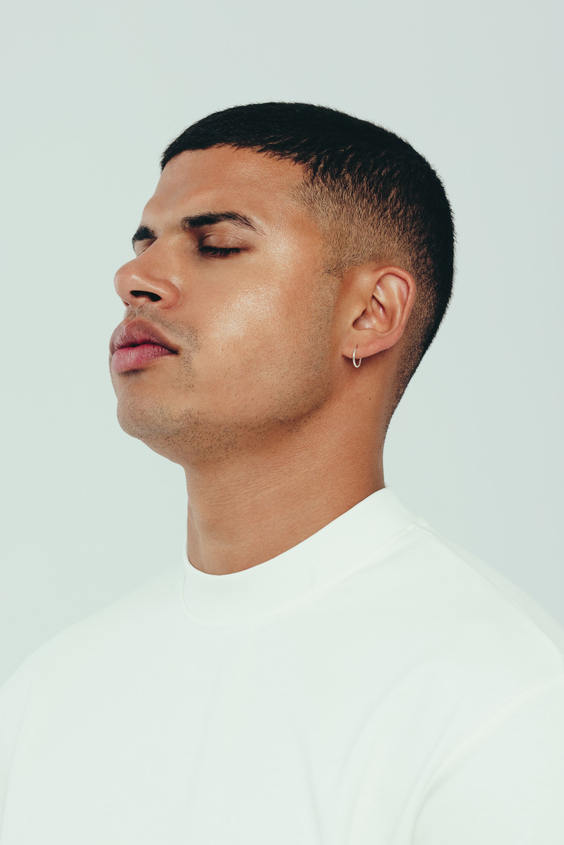 A man wearing a white shirt and earrings is looking down with his eyes closed.