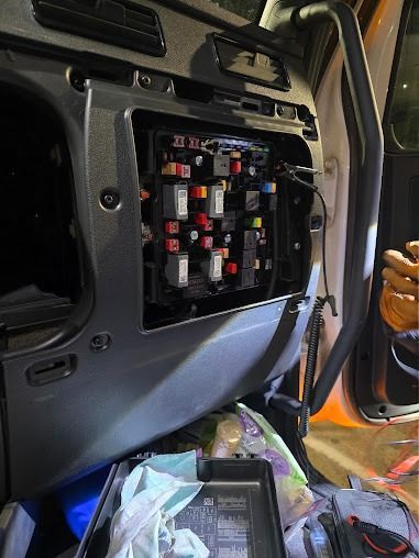 Open vehicle door showing exposed fuse panel and wiring, with tools and bags inside the cabin.