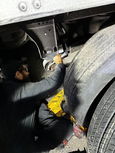 Mechanic working under a vehicle near a large tire, with yellow safety gear visible.