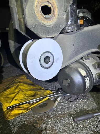 Close-up of a metal car suspension part with a white roller and surrounding tools on a workbench.