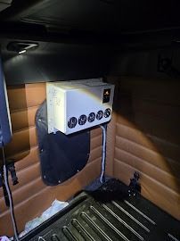 White box mounted inside a car interior above the floor mat, with four black dials and a cable attached.