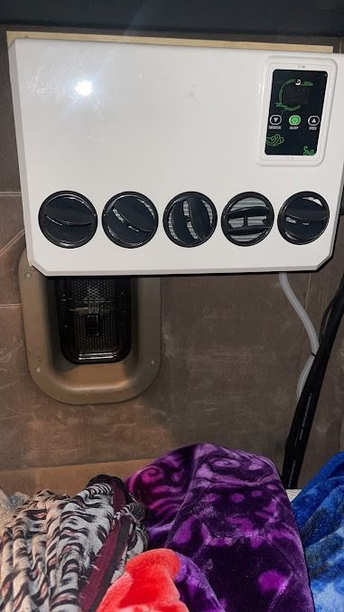 White pet carrier with black vents and a small display, sitting against a wall above a pet bed.
