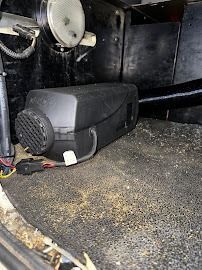 Black heating unit mounted low in a dusty compartment with pipes and a round vent.