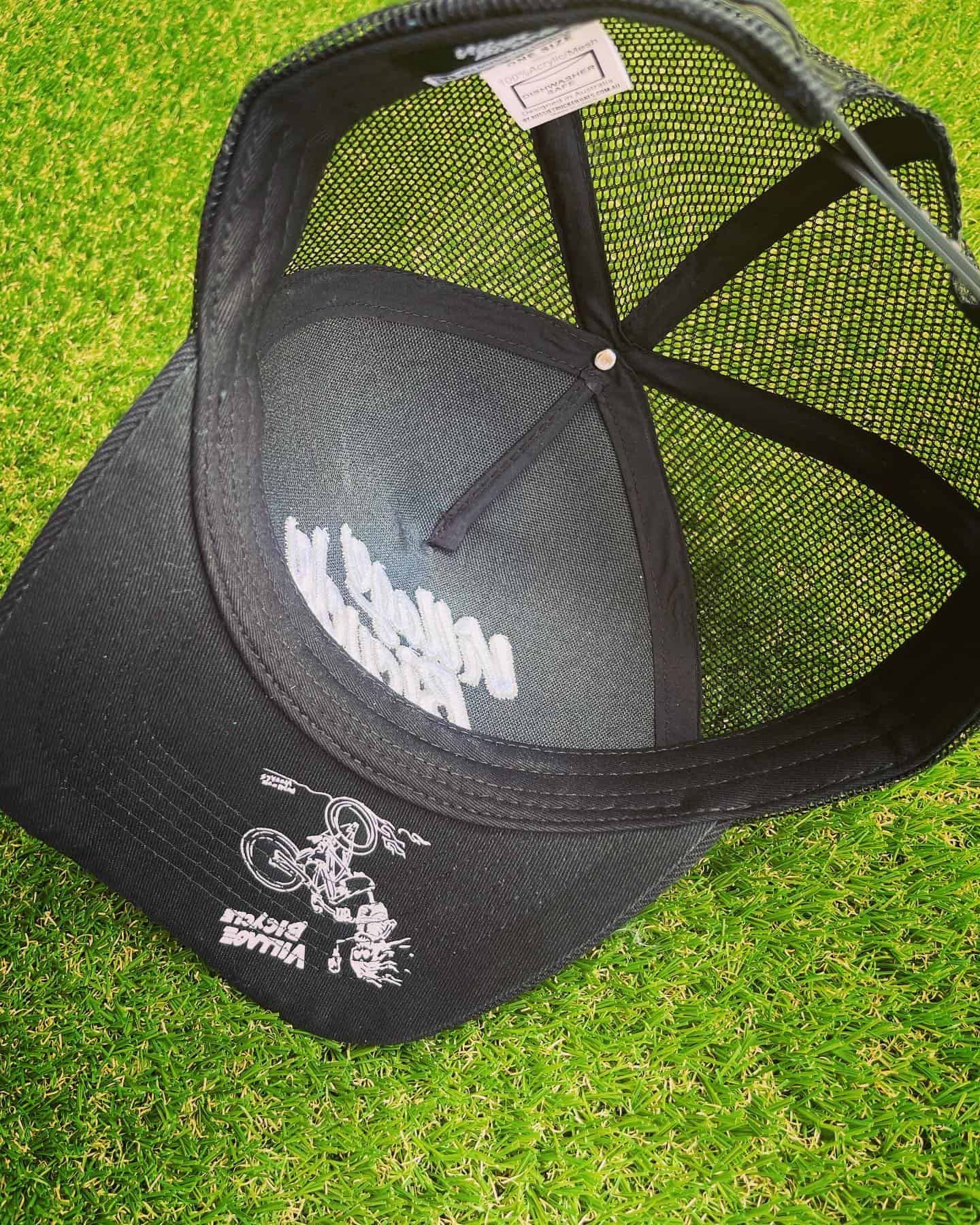 Black trucker hat with mesh back, white printed logo inside against green grass.