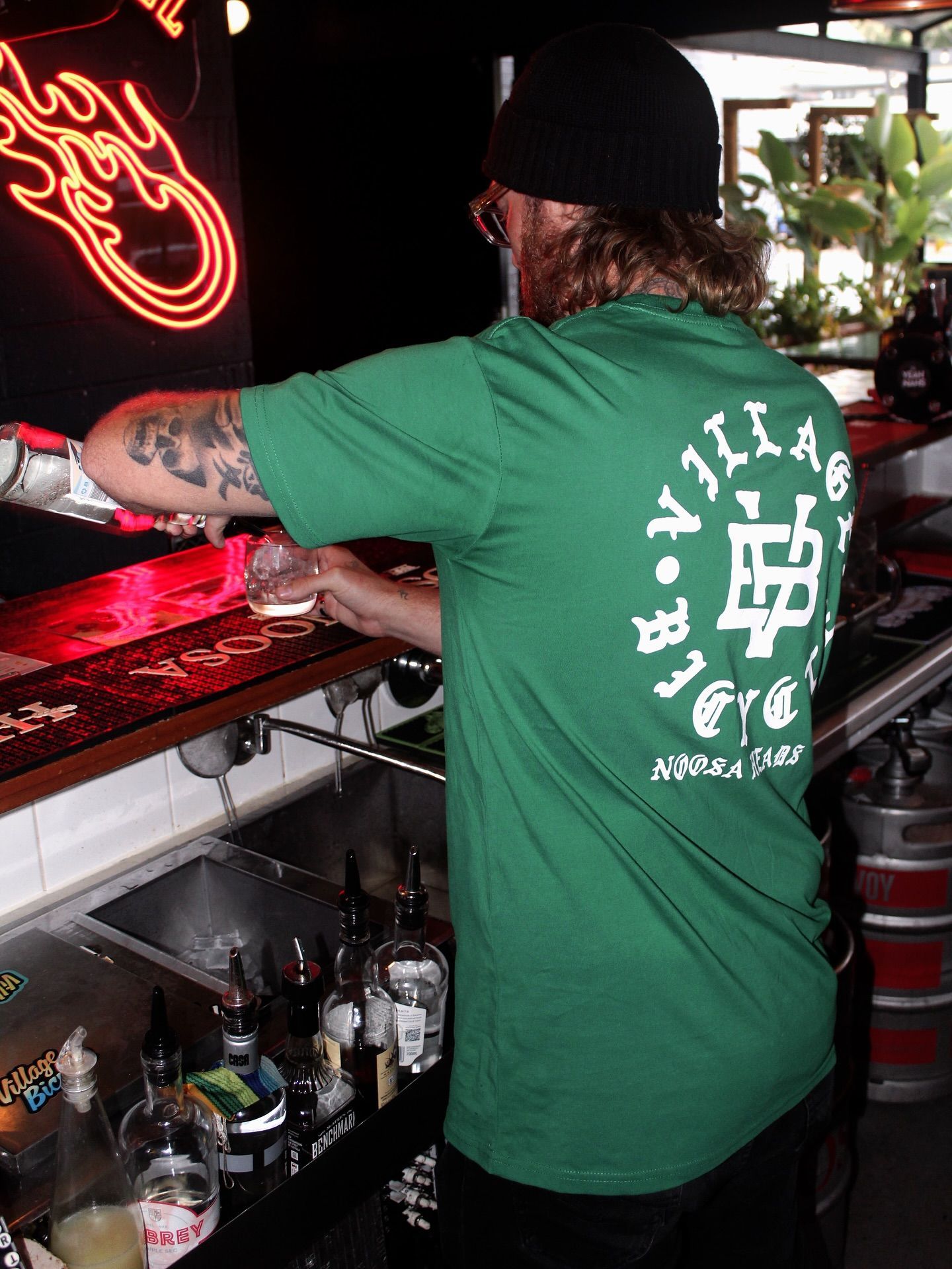Bartender in green shirt pouring drinks behind a bar, flames neon sign in background — Village Bicycle Noosa in Noosa Heads, QLD