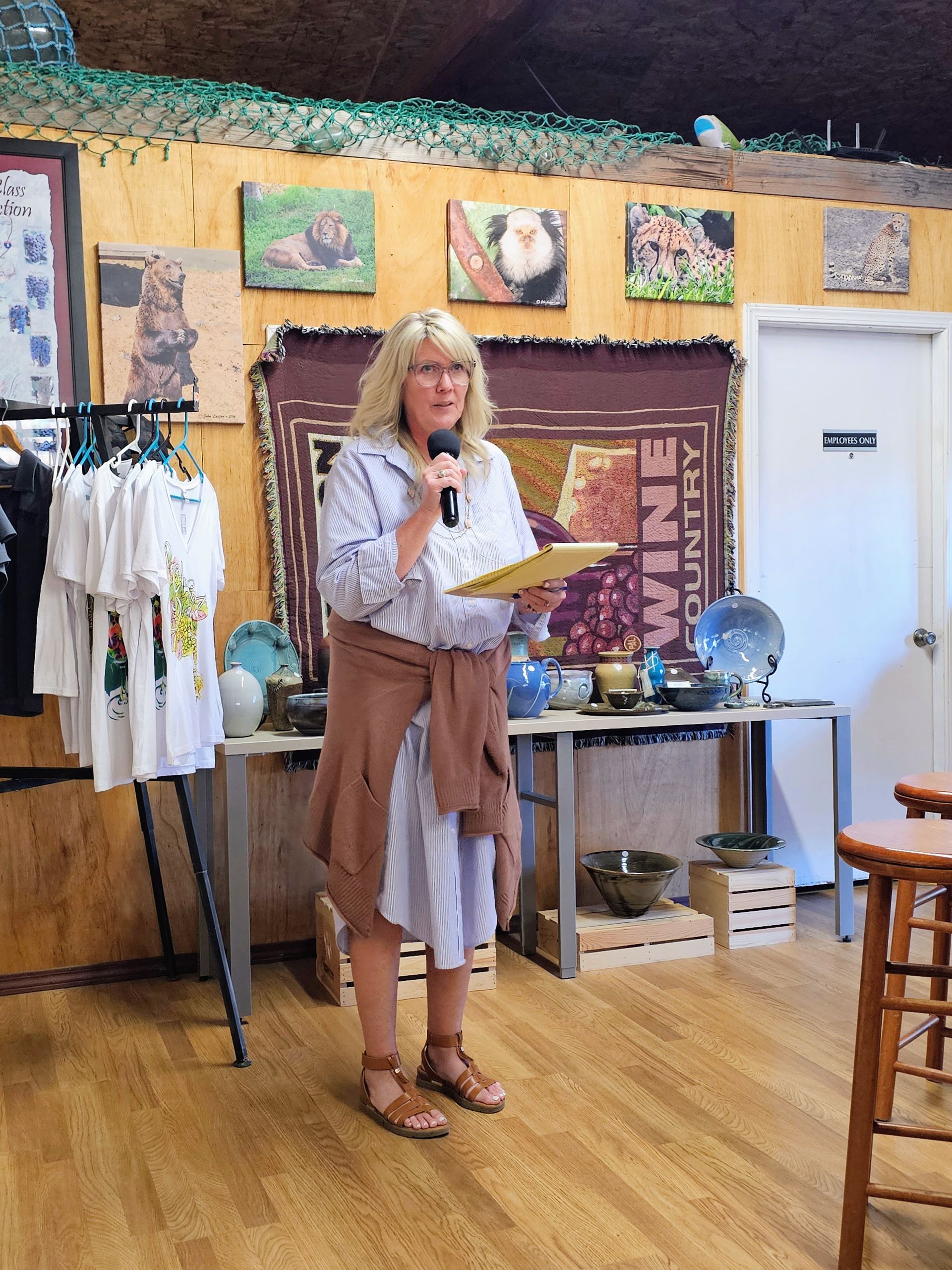 Woman speaking into microphone, holding notes, standing inside a shop with art and merchandise.