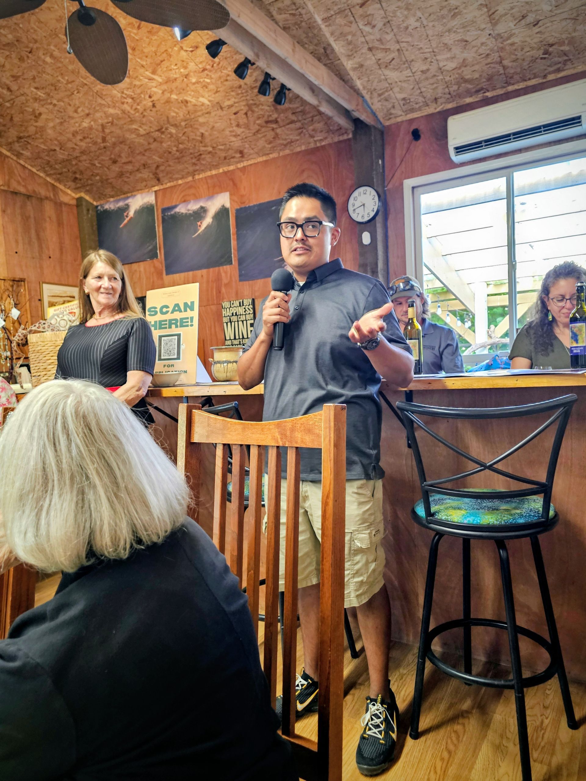 Man speaking into a microphone in a wooden interior, gesturing. Other people listen.