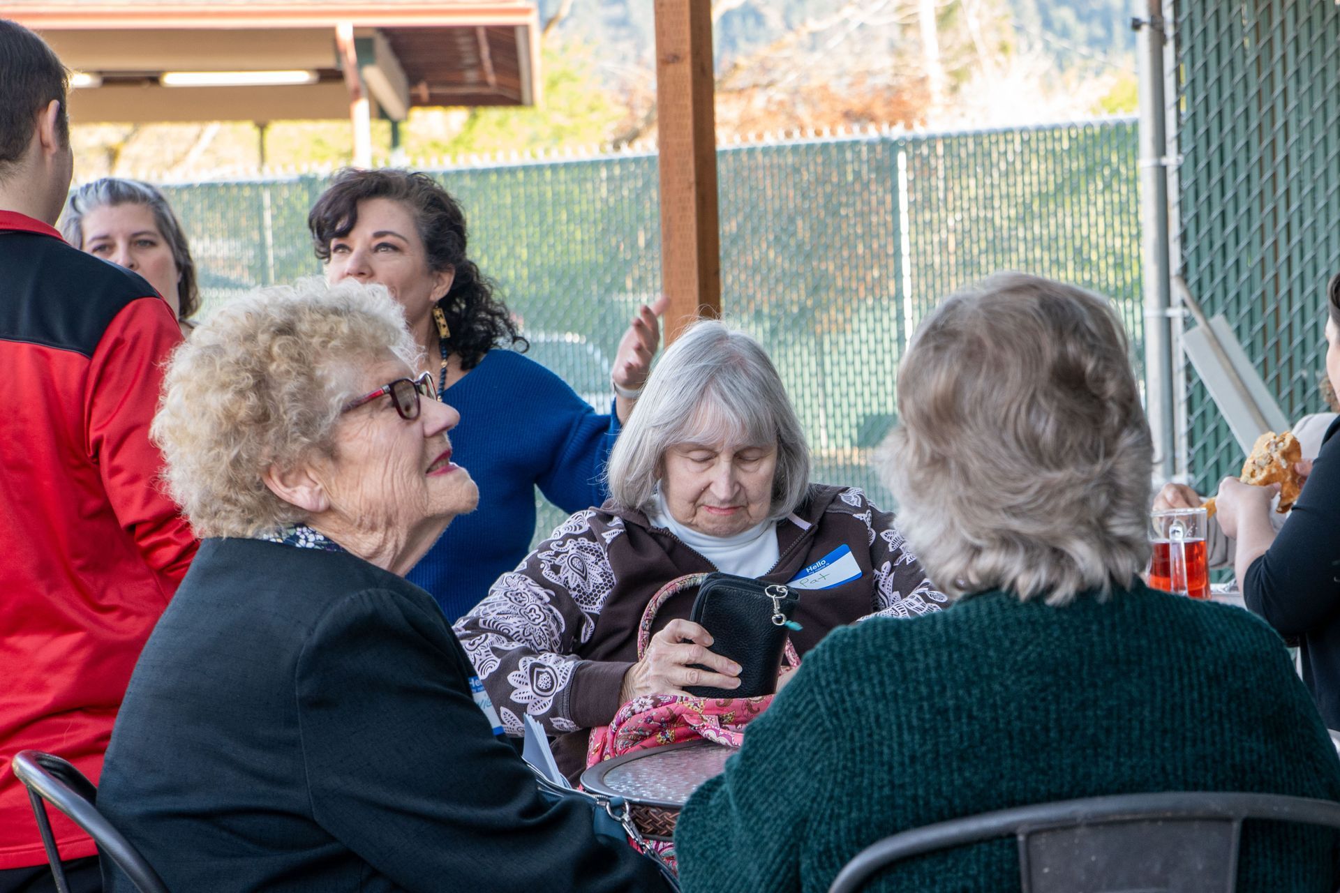Group of people socializing at an outdoor table. One woman is looking at a small purse. Others are talking and smiling.