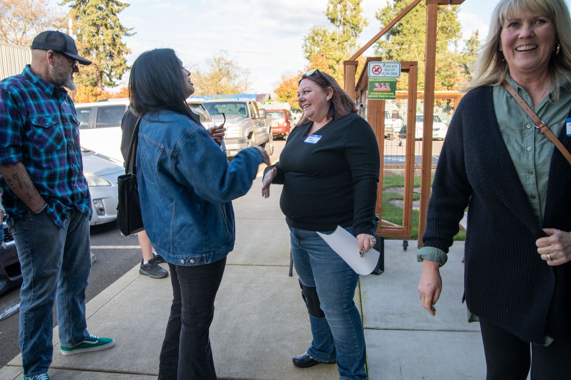 People chatting outdoors, woman laughing. Cars in background, sunny day.