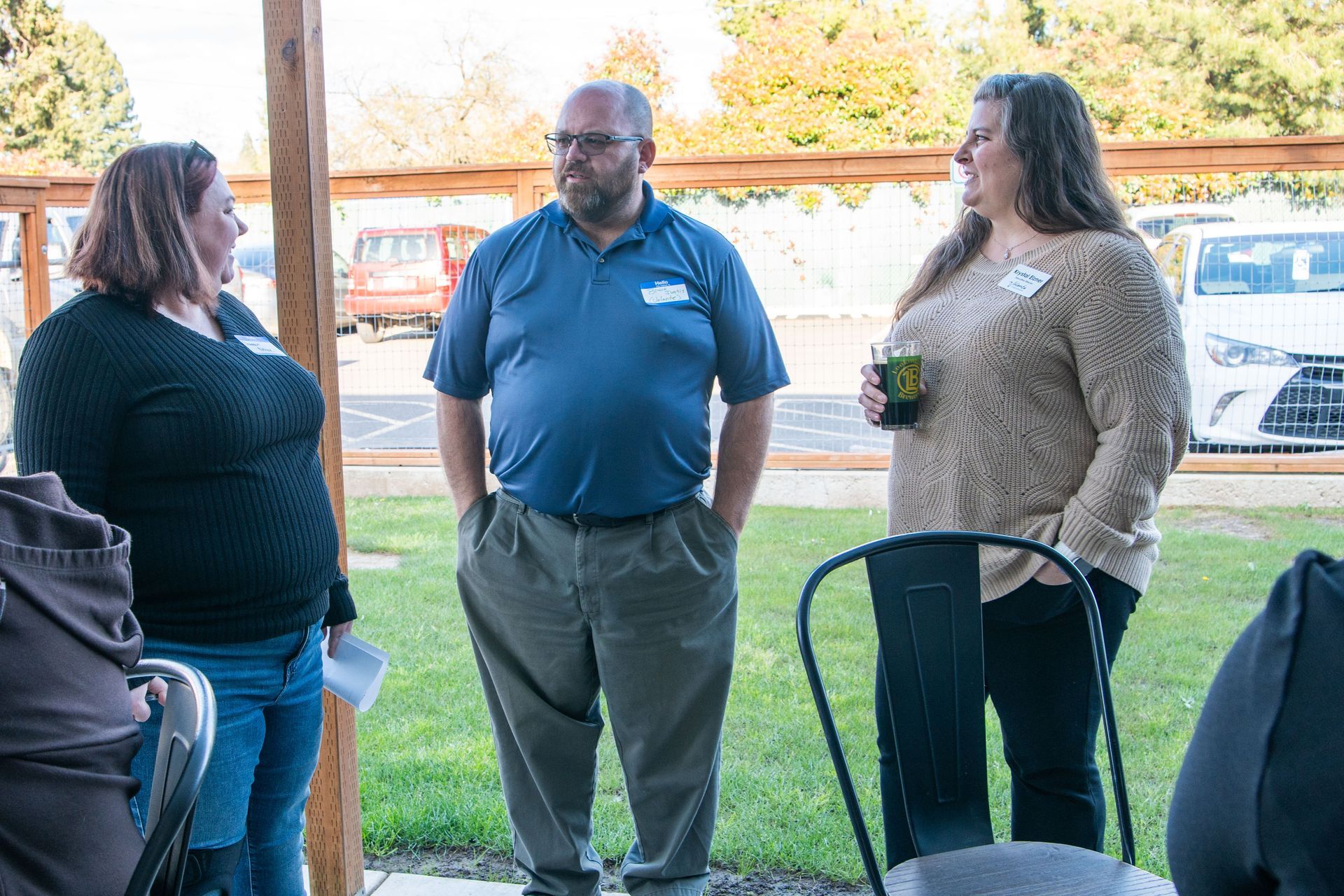 Three people stand outdoors, conversing. Man in blue shirt, two women, one with a drink. Lawn, vehicles in background.