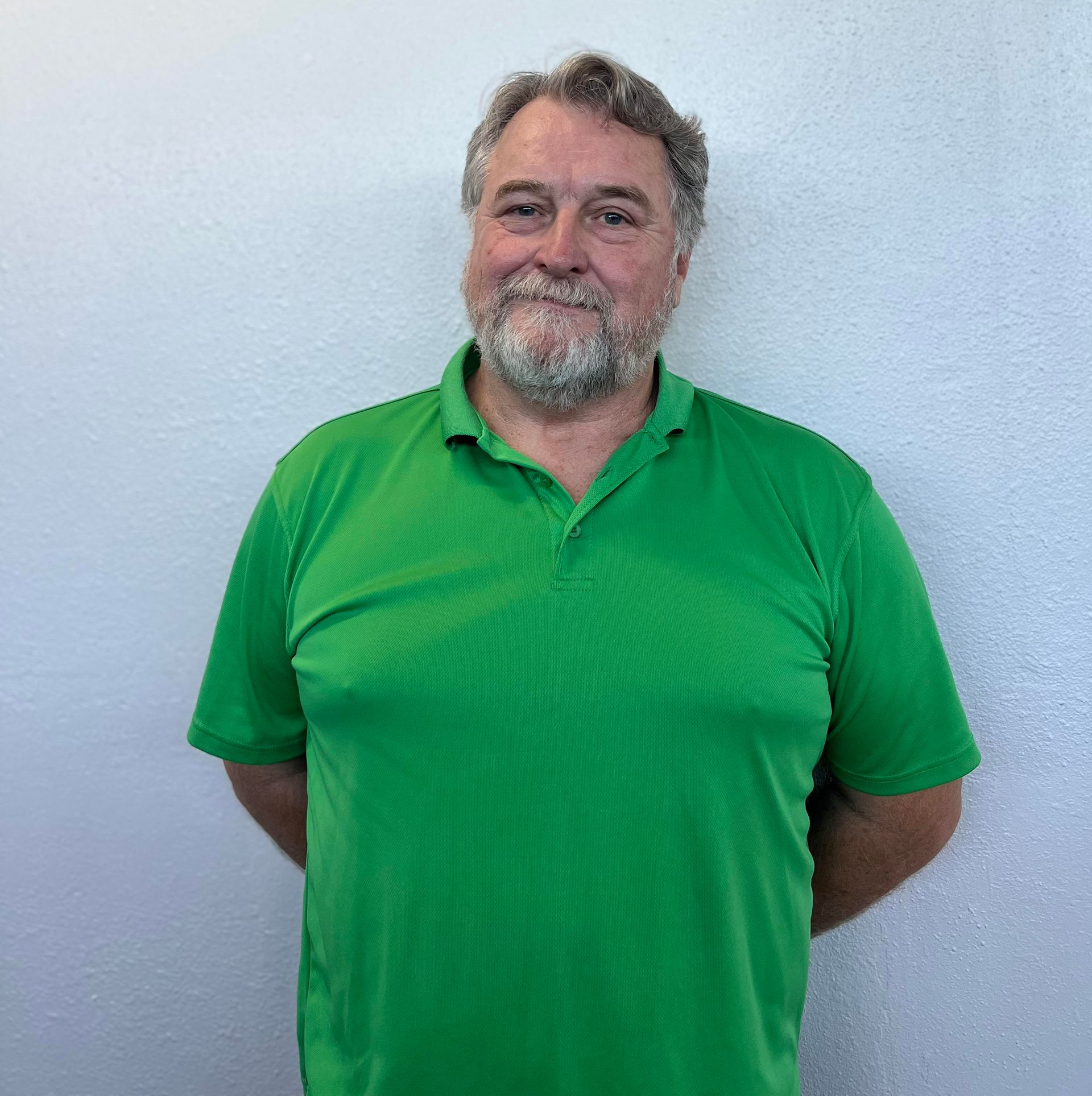 Man with a graying beard, wearing a green polo shirt, standing in front of a white wall, smiling slightly.