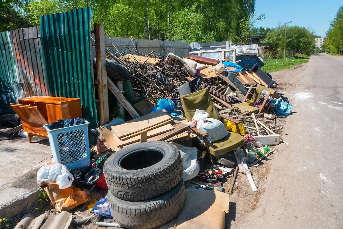 Pile of trash and debris on the side of a road next to a green and gray fence.