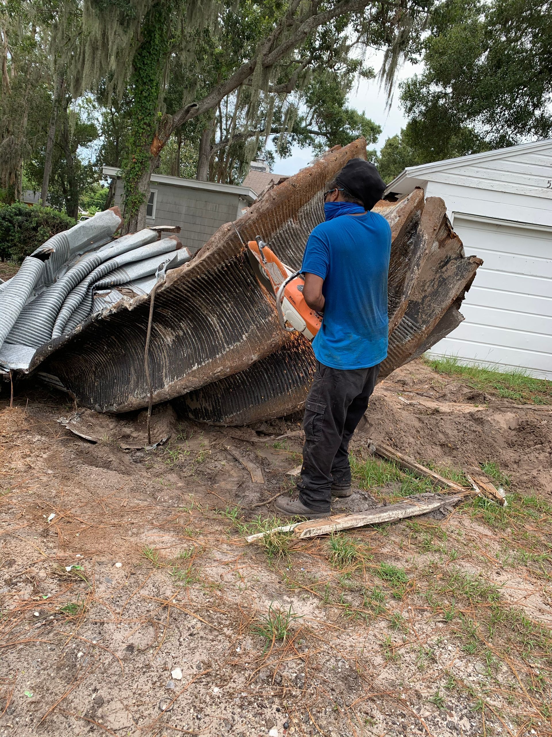 Person in blue shirt using a chainsaw to cut up a large, metal structure on dirt.
