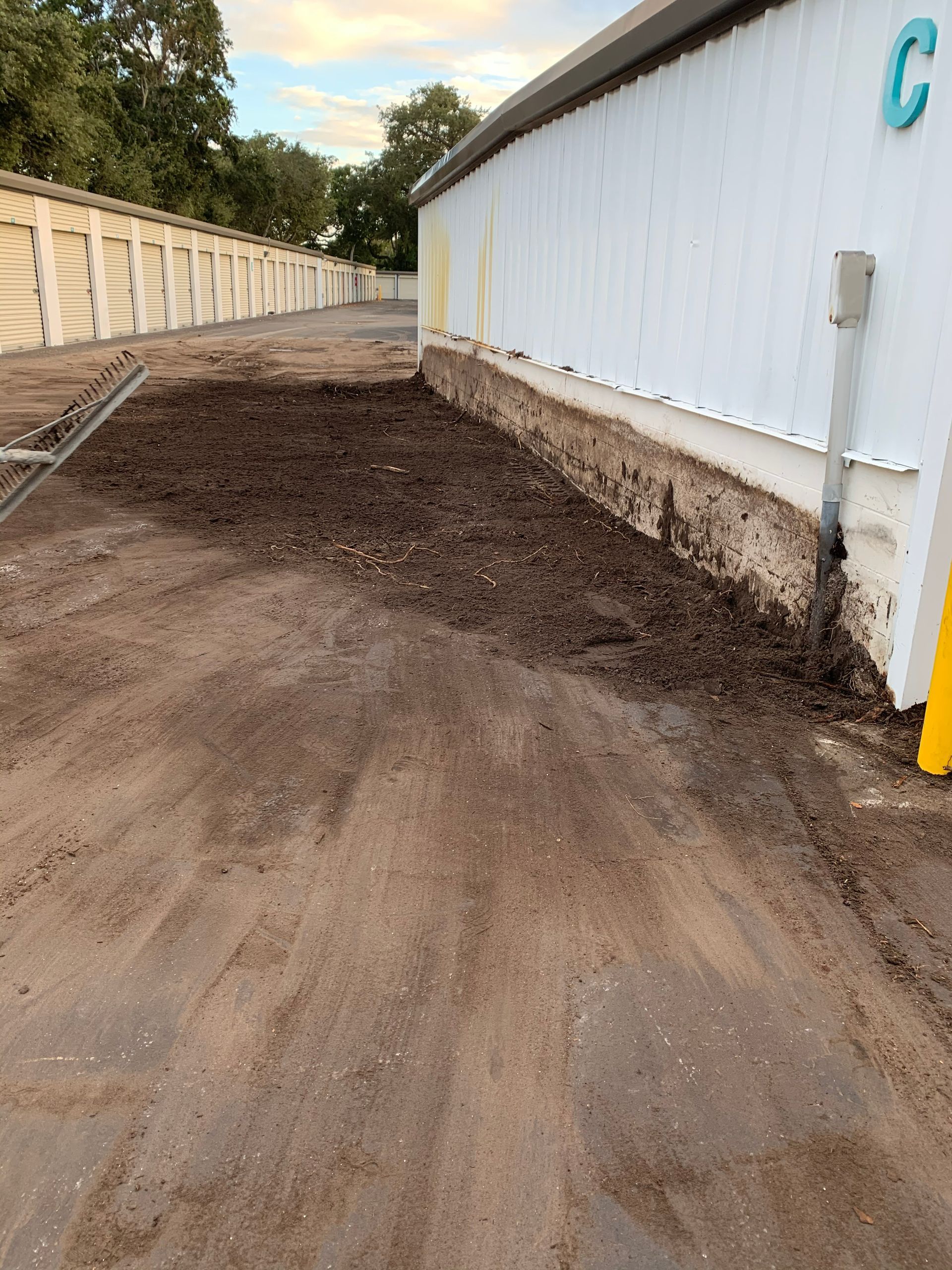 Dirt pathway next to white building with mulch, storage units in background.
