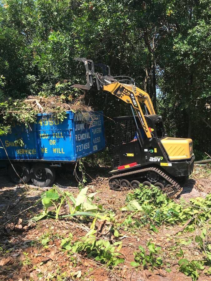 A small yellow tracked excavator loading debris into a blue trailer outdoors.