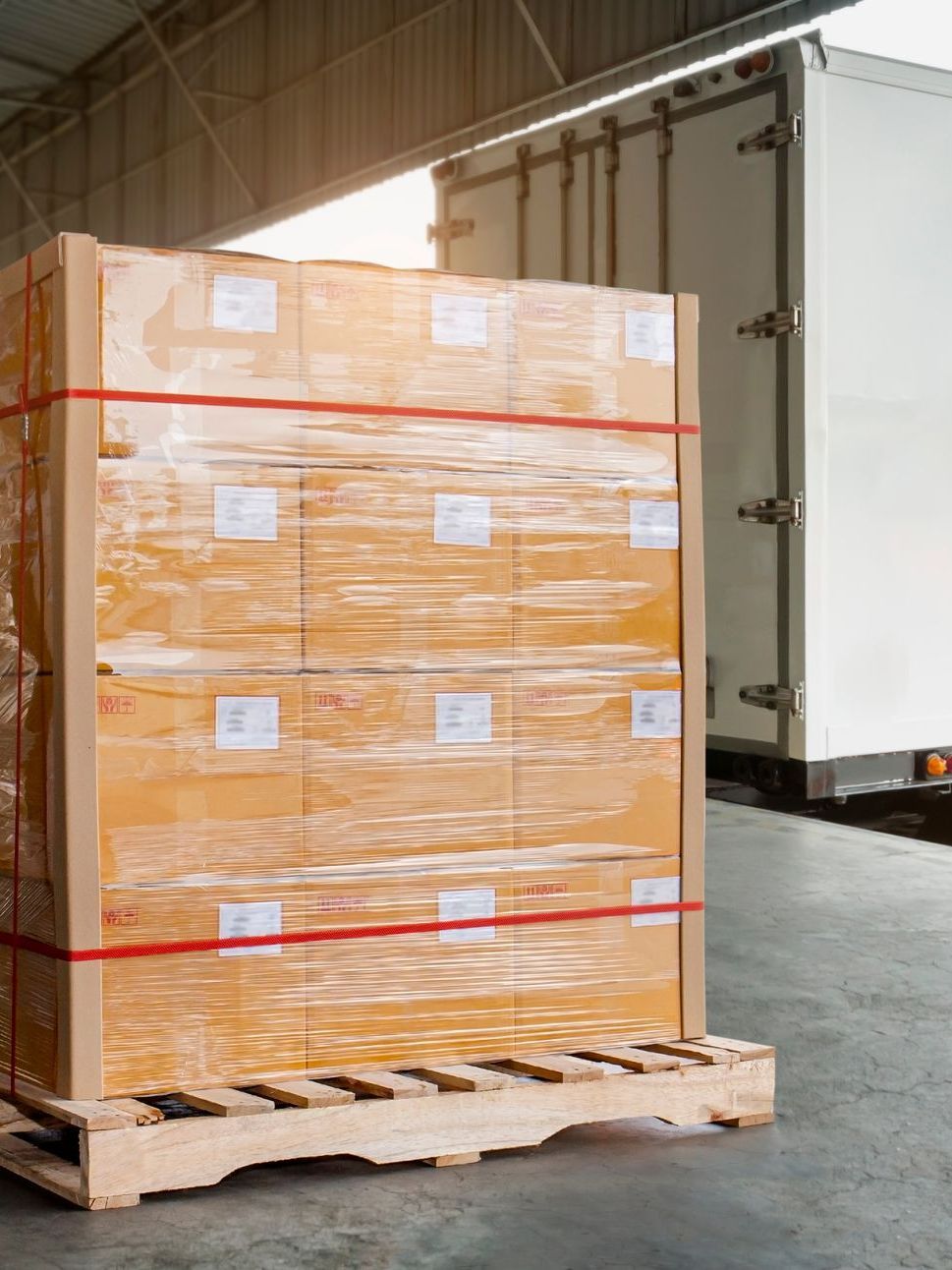 A stack of boxes wrapped in plastic is sitting on a wooden pallet in front of a truck.