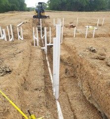 Construction site with buried white PVC pipes in a trench. An excavator is in the background.