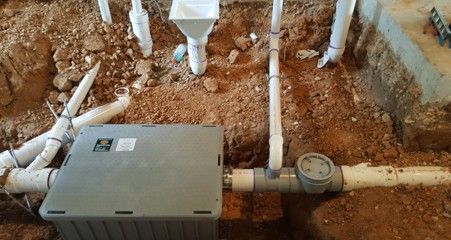 Gray electrical box and water meter connected to white pipes in dirt.