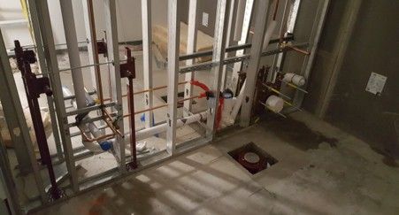 Framed bathroom construction with exposed pipes and plumbing fixtures. Gray walls, concrete floor.