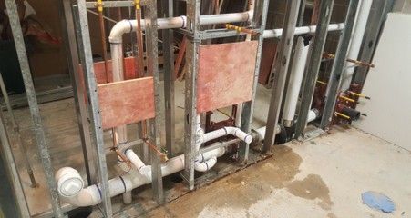 Metal plumbing frame with pipes in progress, plywood squares, concrete floor.