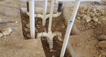 Plumbing pipes in trenches in a construction setting. White PVC pipes with Y-shaped connectors. Brown dirt.