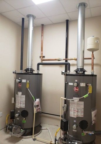 Two gray water heaters with metal chimneys in a utility room. Copper pipes and black pipes connect to the heaters.