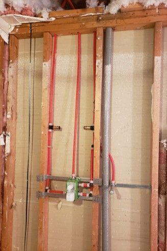 Plumbing rough-in within a wall. Red PEX tubing, green valve, grey vent pipe, against light insulation, wood framing.