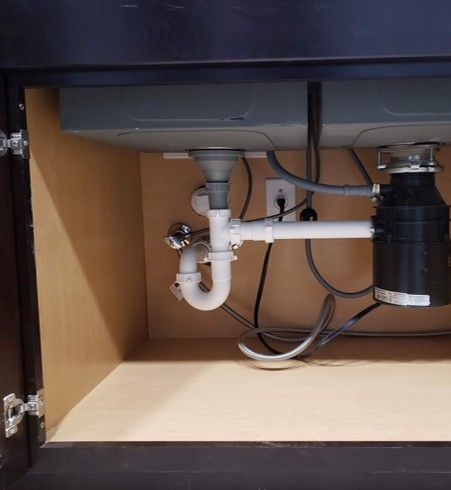 Under-sink plumbing: white pipes, P-trap, garbage disposal, electrical outlet, inside a cabinet.