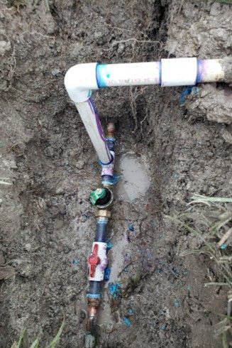 Underground irrigation system components: PVC pipes, valves, and fittings in a dirt trench.