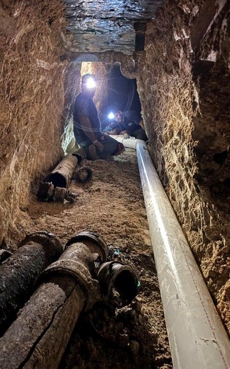 Person in tunnel with headlamp, inspecting pipes. Brown walls, white pipe, and dirty metal pipes.