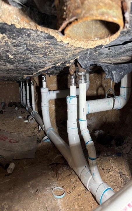 White PVC pipes installed under a building, branching from metal fixtures. Dark, earthen ground.