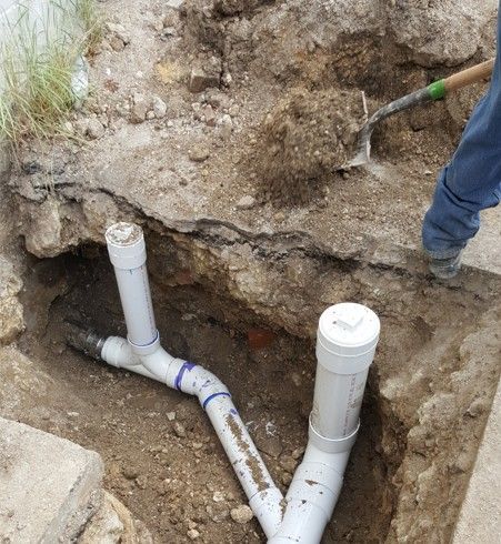 A person digging in a trench to access PVC pipes; the white pipes branch and turn.