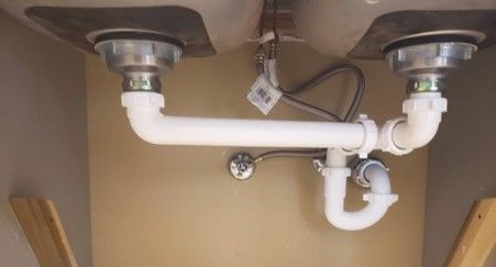 Under-sink plumbing with white pipes, two sink drains, and a U-shaped trap in a beige cabinet.