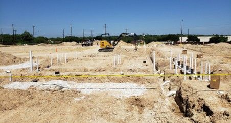 Construction site with excavator, trenches, and pipes laid out in the dirt under a blue sky.