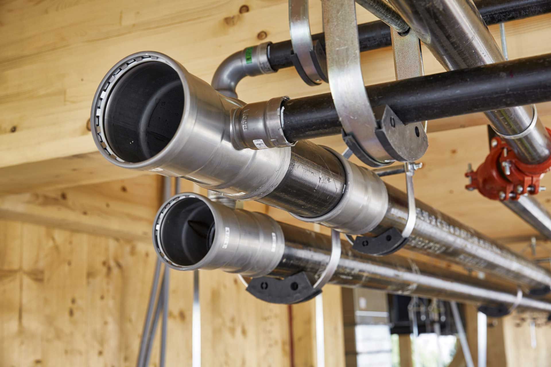 Pipes suspended from wooden ceiling, various sizes and materials, with metal brackets and fittings.