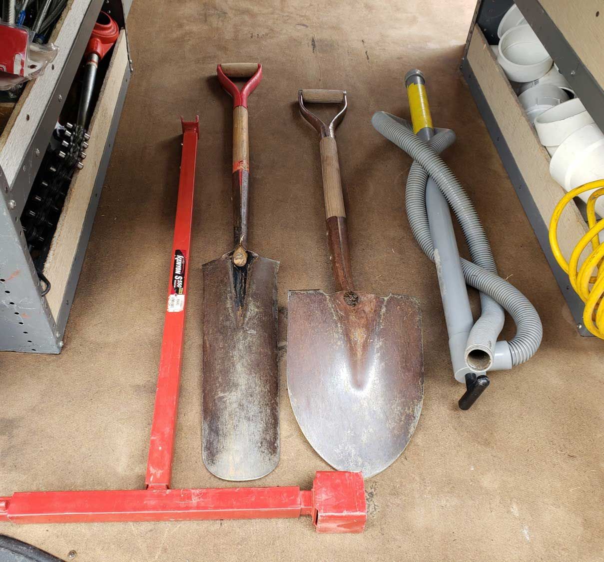 Two shovels, a red tool, and a vacuum hose in a tool compartment.