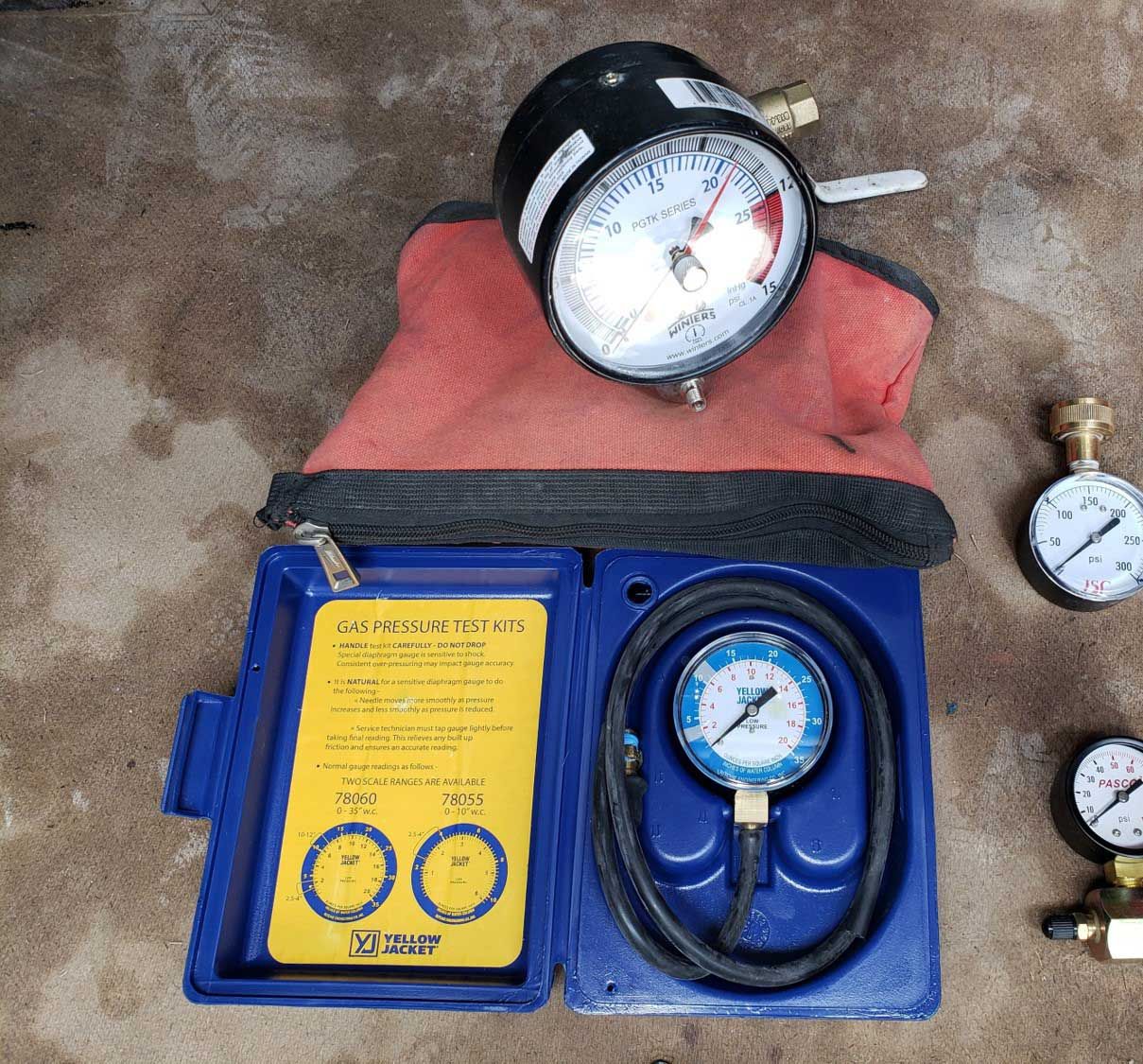 Open blue case with a pressure gauge, a gauge with a red covering, and two other gauges on concrete.
