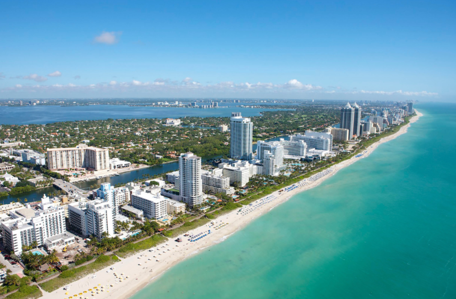 Aerial view of a coastal city with high-rise buildings, white sandy beach, and turquoise ocean under a clear blue sky