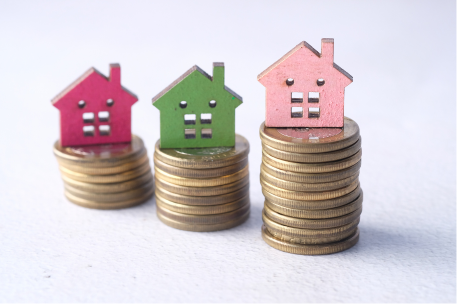 Three small house figurines in pink, green, and peach stacked on coin piles, symbolizing home savings or property value