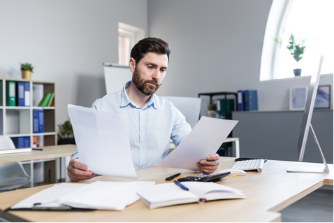 Man reviewing documents at a desk in a bright office, with papers, folders, and a computer monitor nearby.