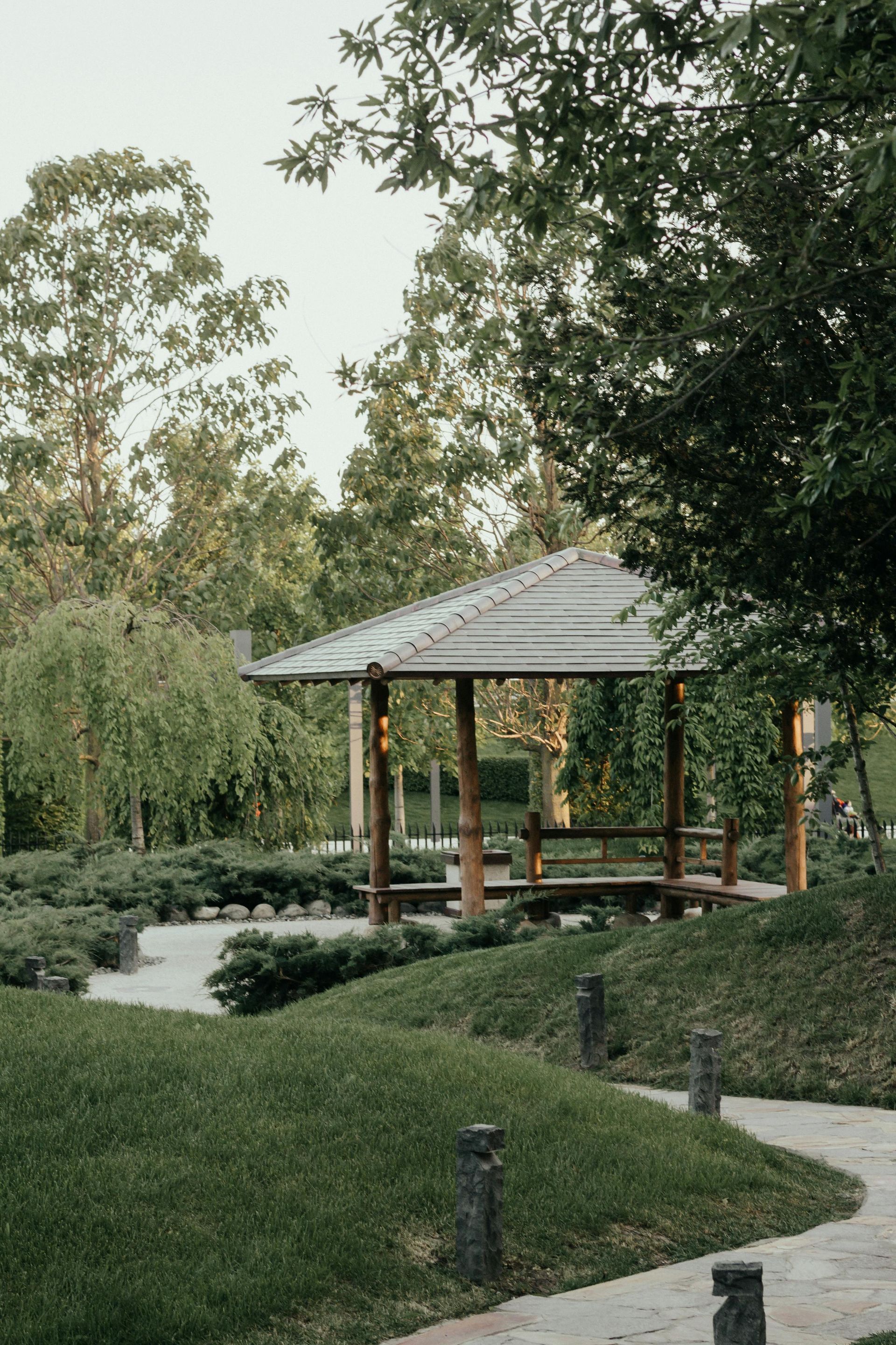 Park gazebo surrounded by greenery with a winding path and small lights at dusk