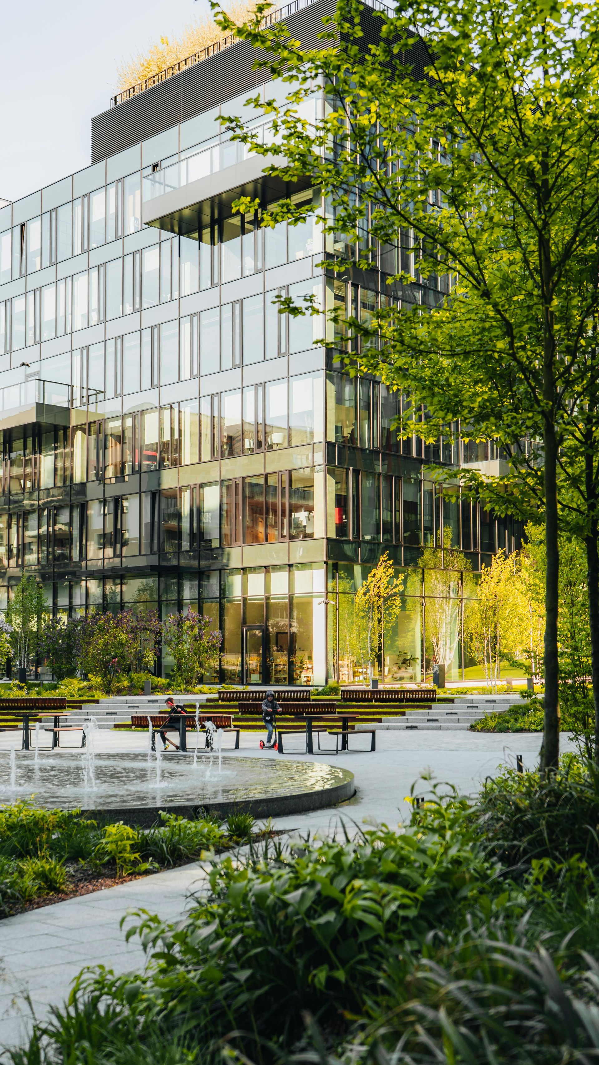 Modern glass office building beside a landscaped courtyard with benches and lush green trees