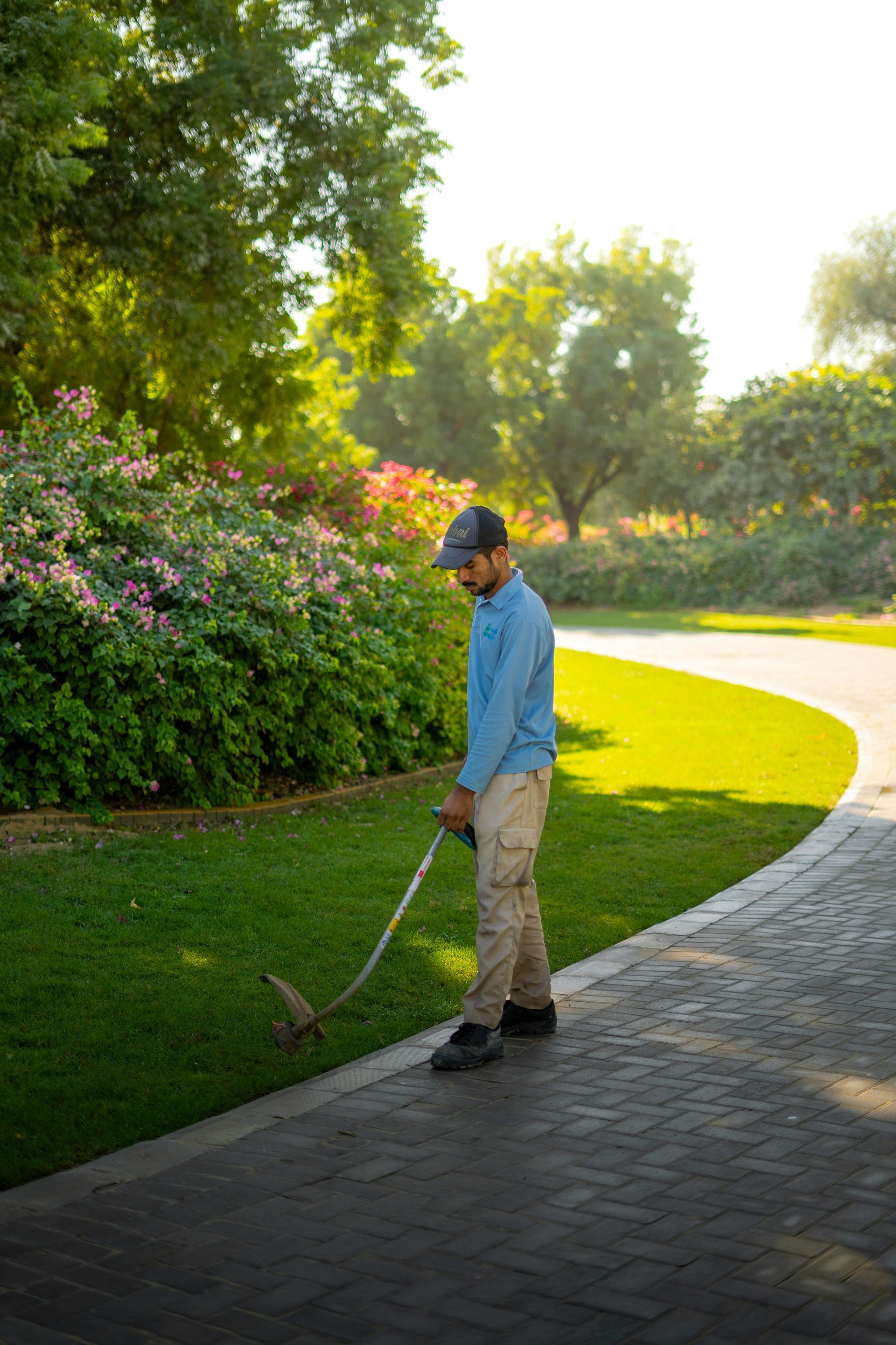 Man using a golf club on a paved path beside a green lawn and flowering bushes in a park