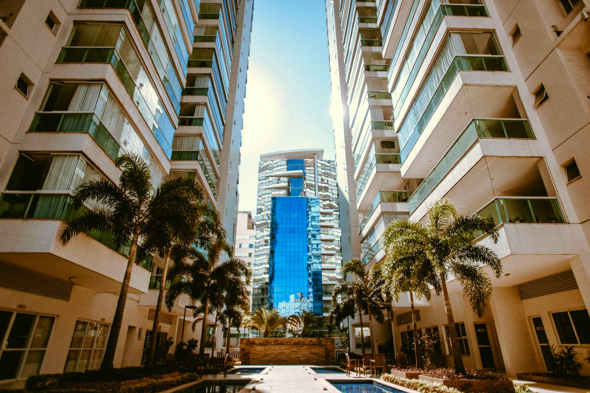 Sunlit city courtyard framed by tall white apartment towers and palm trees, with a blue glass building in the center