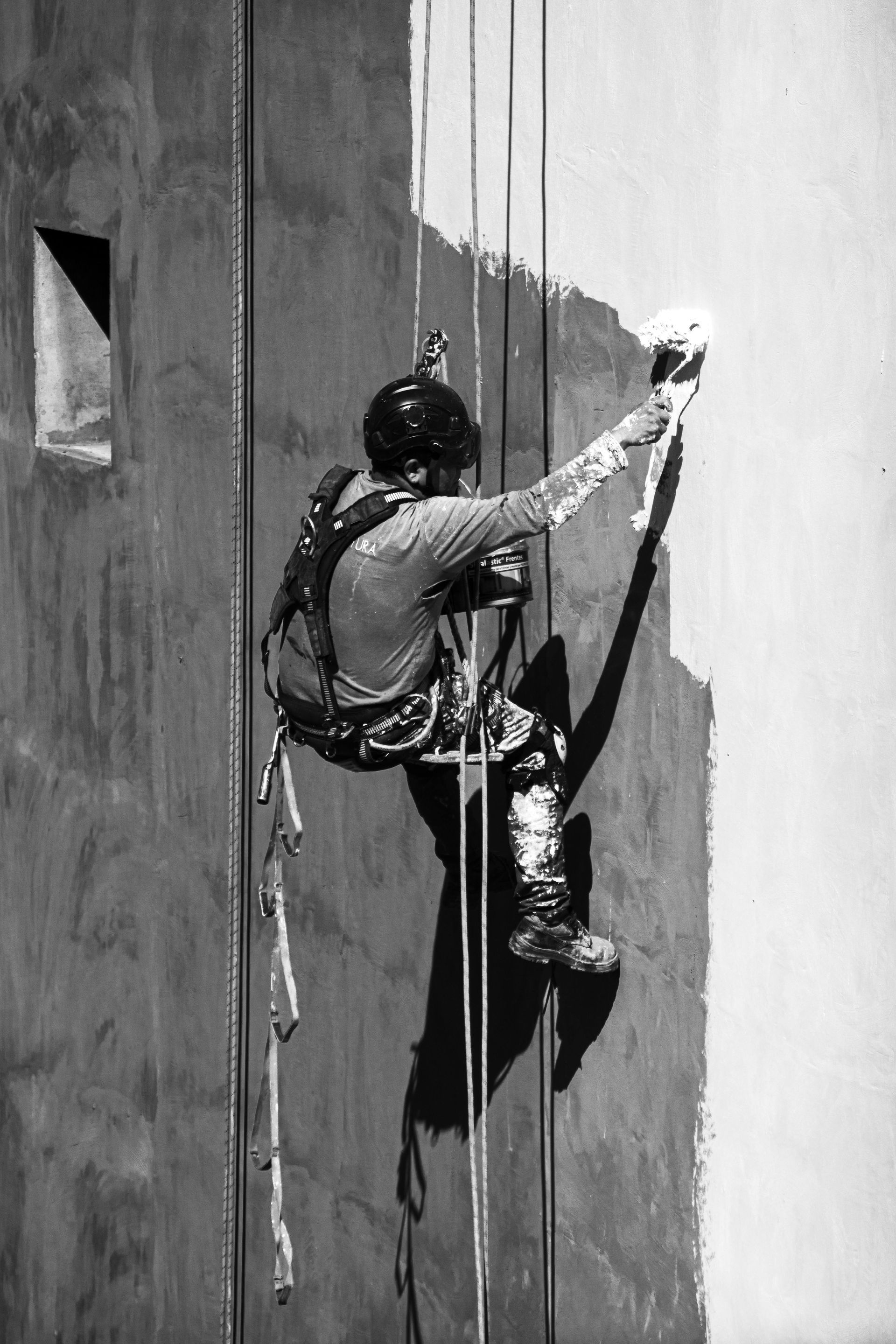 Worker suspended by ropes painting a tall wall, with a sharp vertical split of dark and light paint.