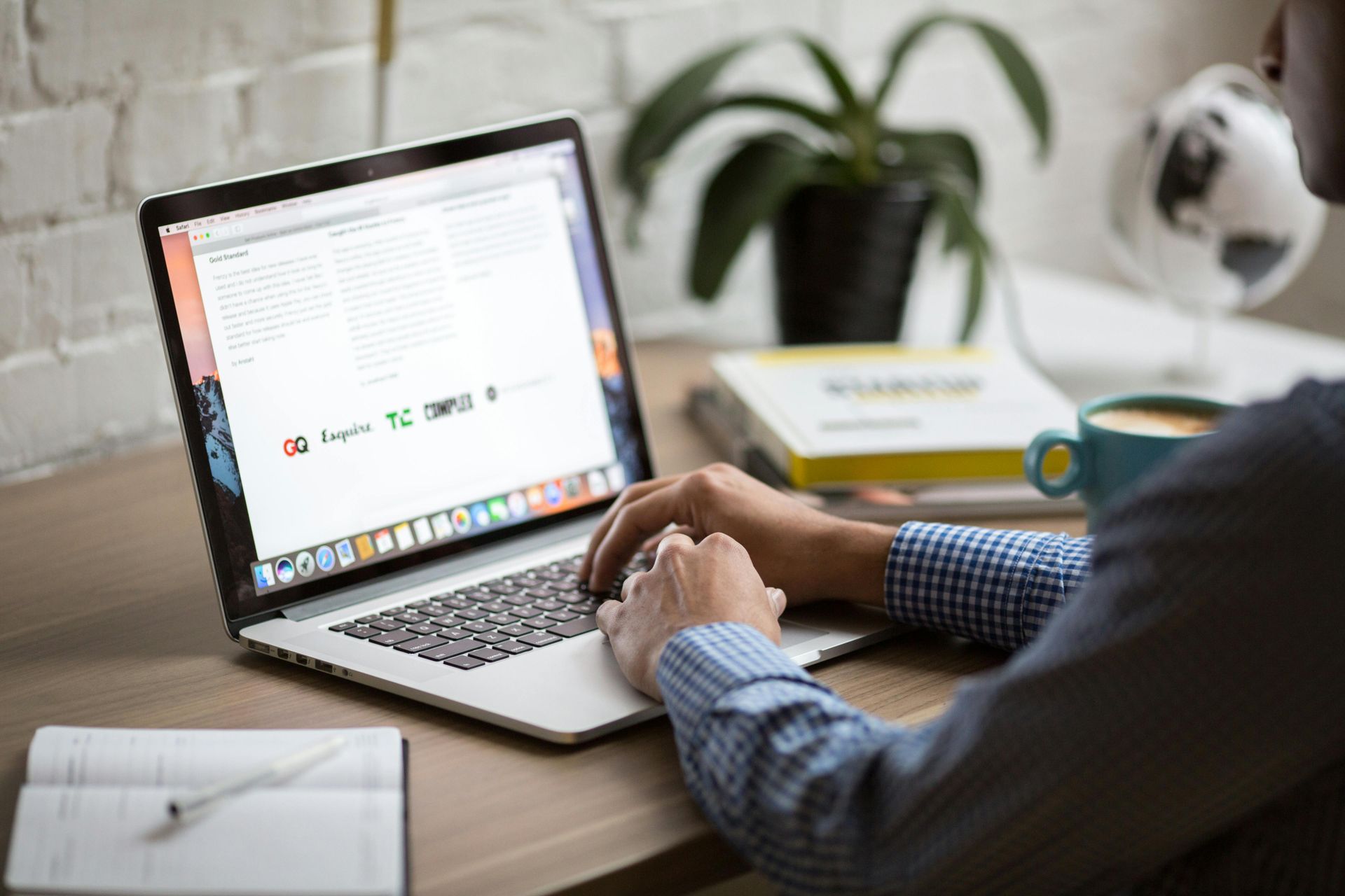 Person typing on a laptop at a desk with a notebook and plant in the background