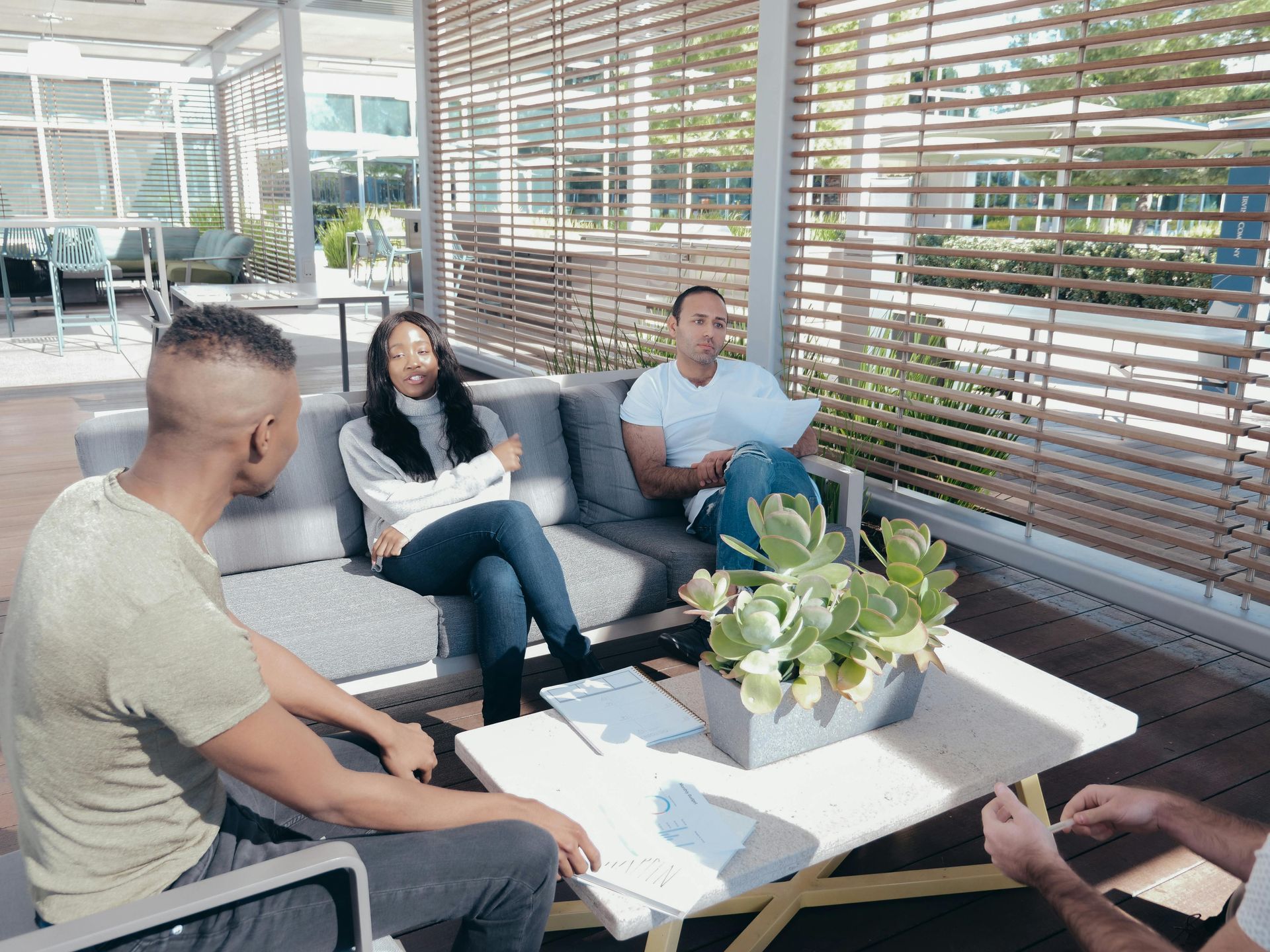 Three people chatting on a patio around a white coffee table with a potted plant.