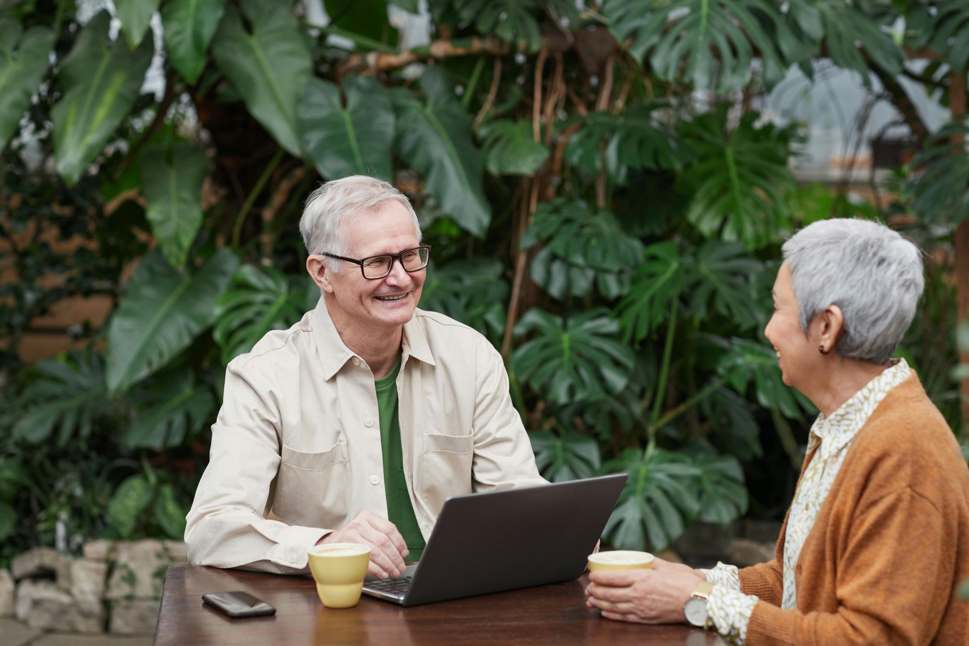 Two people smiling and chatting at an outdoor table with a laptop and drinks, surrounded by greenery