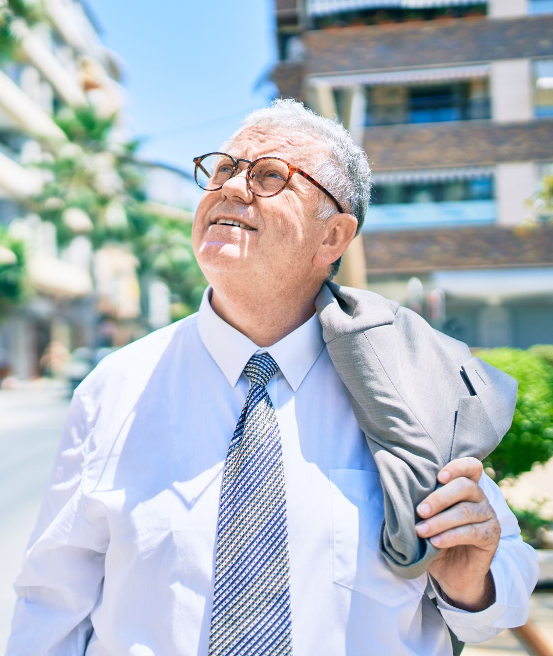 Man in a white shirt and tie smiles upward outdoors, holding a gray jacket over his shoulder.